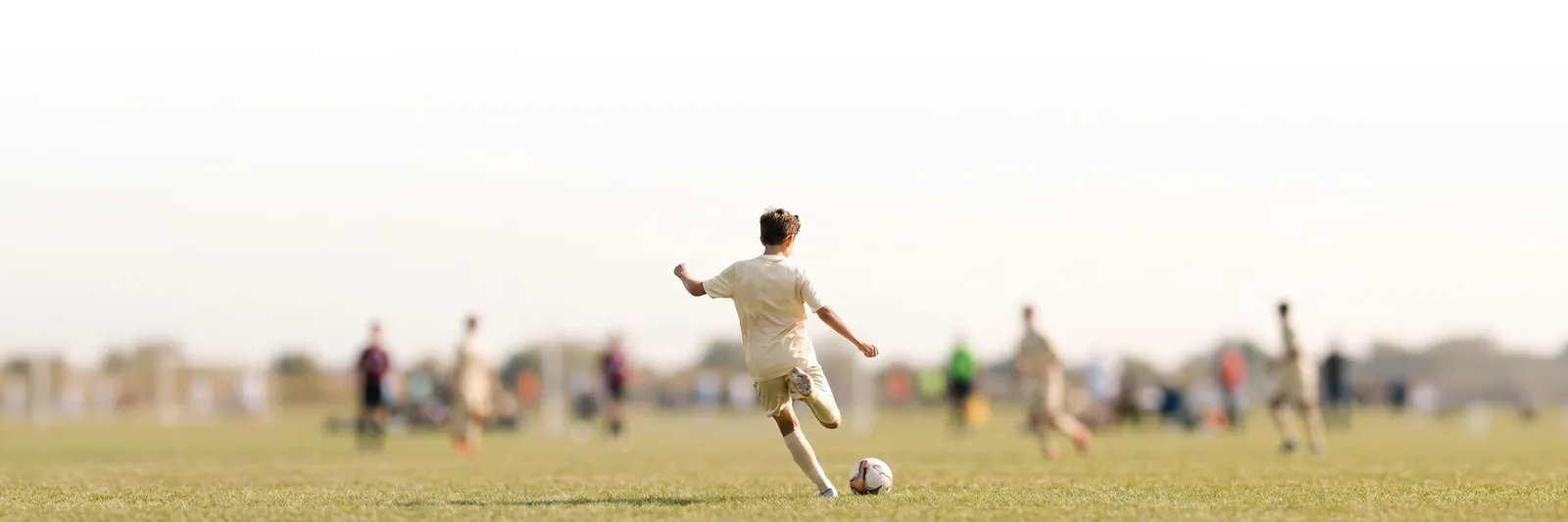 A child running up to strike a football on a wide grass pitch, with other players and a sideline out of focus behind